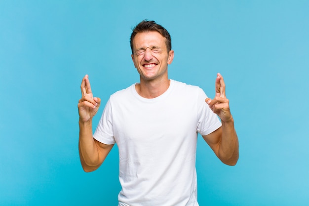 Young handsome man smiling and anxiously crossing both fingers, feeling worried and wishing or hoping for good luck