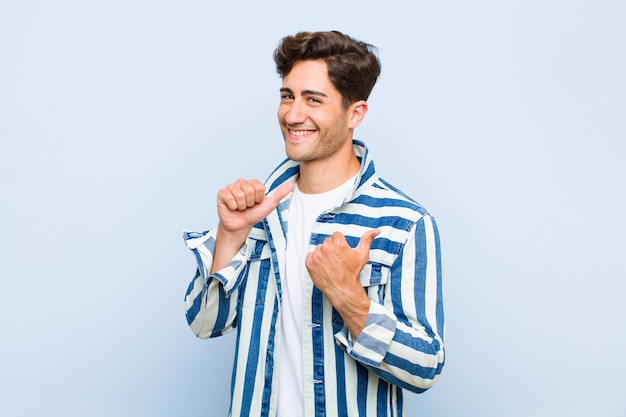 Young handsome man smiling cheerfully and casually pointing to copy space on the side, feeling happy and satisfied against blue background