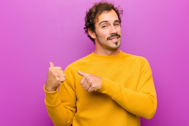 Young handsome man smiling cheerfully and casually pointing to copy space on the side, feeling happy and satisfied over purple wall