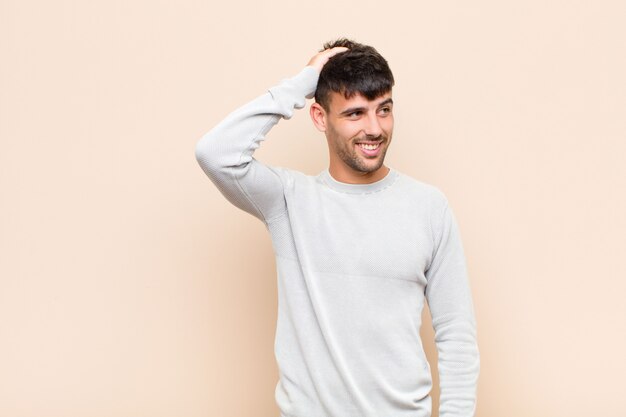 Young handsome man smiling cheerfully and casually, taking hand to head with a positive, happy and confident look against warm wall