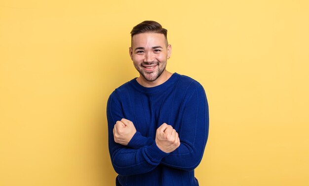 Young handsome man smiling cheerfully and celebrating, with fists clenched and arms crossed, feeling happy and positive