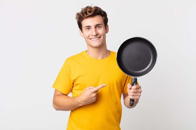 元気に笑って、幸せを感じて、横を指して、調理鍋を持っている若いハンサムな男