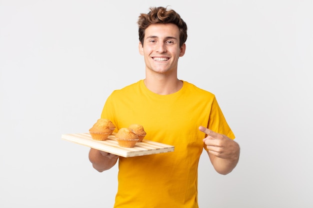 Young handsome man smiling cheerfully, feeling happy and pointing to the side holding a muffins tray