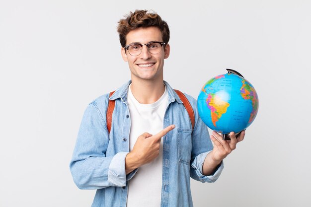 元気に笑って、幸せを感じて、横を指している若いハンサムな男。世界の地球地図を持っている学生