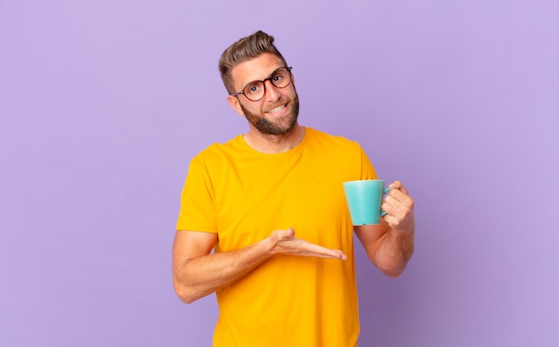 Giovane uomo bello che sorride allegramente, si sente felice e mostra un concetto. e con in mano una tazza di caffè