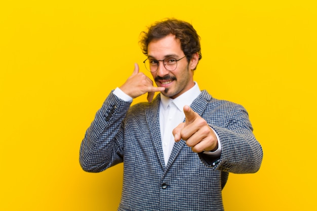 Young handsome man smiling cheerfully and pointing to camera while making a call you later gesture, talking on phone against orange wall