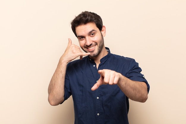 Young handsome man smiling cheerfully and pointing while making a call you later gesture, talking on phone