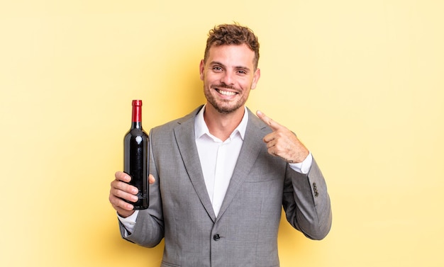 Young handsome man smiling confidently pointing to own broad smile. bottle of wine concept