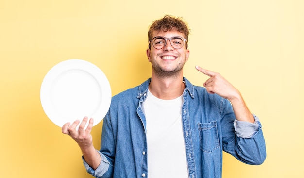 Young handsome man smiling confidently pointing to own broad smile. empty dish concept