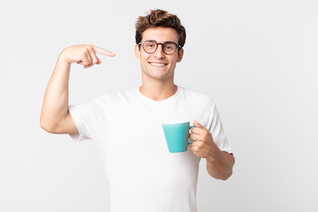 自信を持って笑顔の若いハンサムな男は、自分の広い笑顔を指して、コーヒーカップを持っています