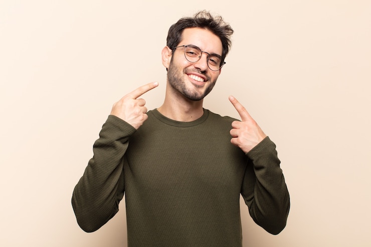 Photo young handsome man smiling confidently pointing to own broad smile, positive, relaxed, satisfied attitude