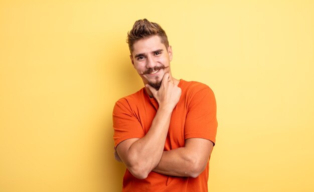 Young handsome man smiling enjoying life feeling happy friendly satisfied and carefree with hand on chin strange moustache concept