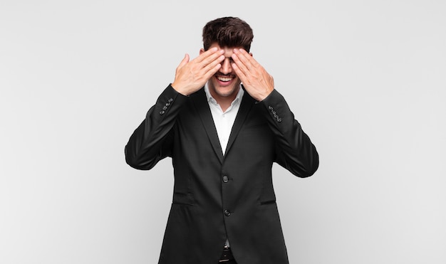 Young handsome man smiling and feeling happy, covering eyes with both hands and waiting for unbelievable surprise