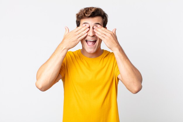 Young handsome man smiling and feeling happy, covering eyes with both hands and waiting for unbelievable surprise