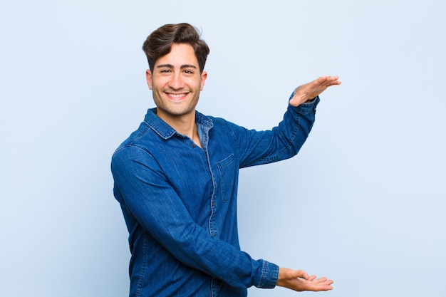 Young handsome man smiling, feeling happy, positive and satisfied, holding or showing object or concept on copy space against blue background