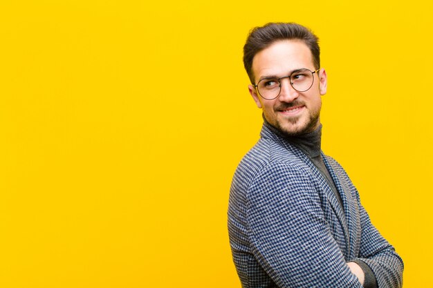 Young handsome man smiling gleefully, feeling happy, satisfied and relaxed, with crossed arms and looking to the side against orange wall