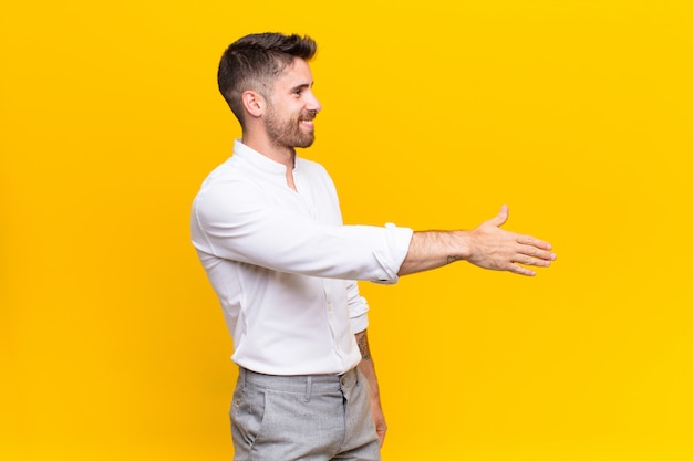 Young handsome man smiling, greeting you and offering a hand shake to close a successful deal, cooperation concept against flat color wall