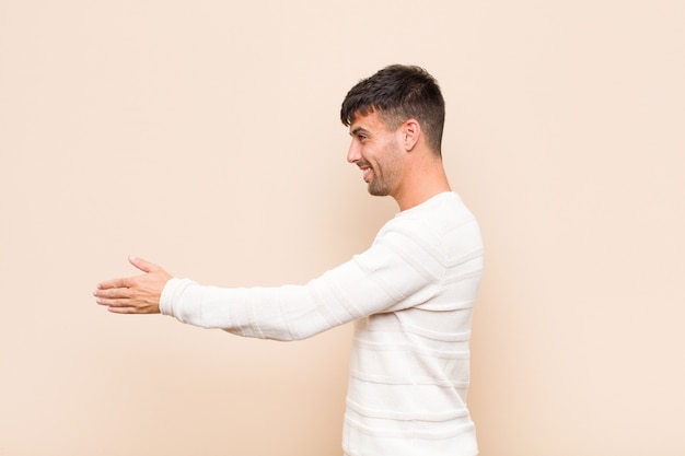 Photo young handsome man smiling, greeting you and offering a hand shake to close a successful deal, cooperation concept over warm wall