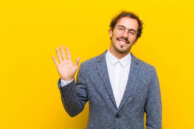 Young handsome man smiling happily and cheerfully, waving hand, welcoming and greeting you, or saying goodbye against orange wall