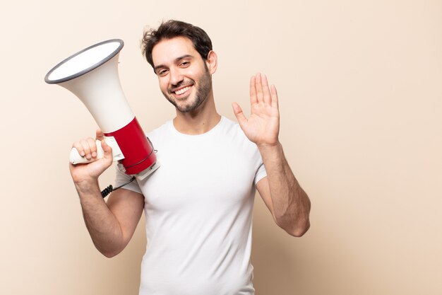 Young handsome man smiling happily and cheerfully, waving hand, welcoming and greeting you, or saying goodbye