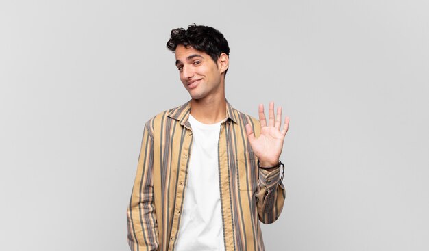 young handsome man smiling happily and cheerfully, waving hand, welcoming and greeting you, or saying goodbye