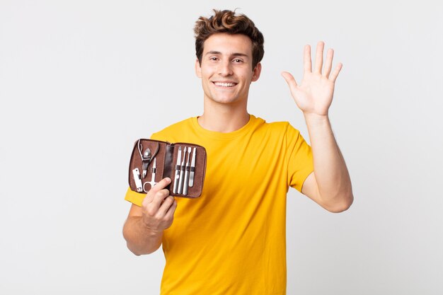 Young handsome man smiling happily, waving hand, welcoming and greeting you and holding a nails tools case