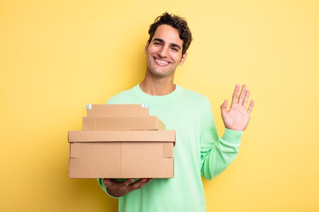 Young handsome man smiling happily, waving hand, welcoming and greeting you. package boxes