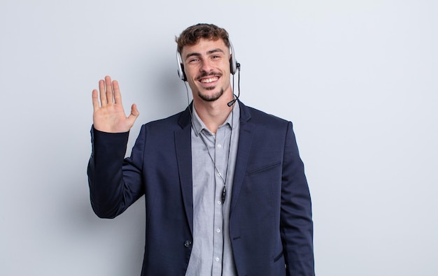 Photo young handsome man smiling happily, waving hand, welcoming and greeting you. telemarketer concept