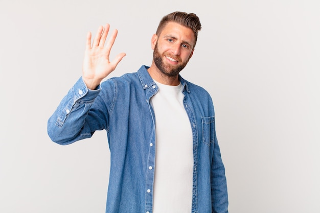 Young handsome man smiling happily, waving hand, welcoming and greeting you