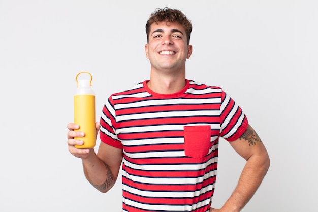 Young handsome man smiling happily with a hand on hip and confident and holding a coffee thermos