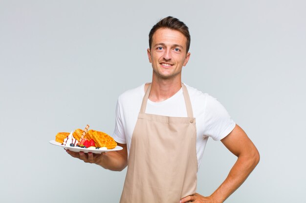 Young handsome man smiling happily with a hand on hip and confident, positive, proud and friendly attitude