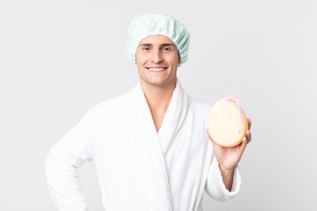 Young handsome man smiling happily with a hand on hip and confident with bathrobe, shower cap and a sponge