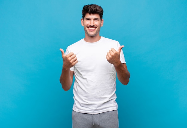 Young handsome man smiling joyfully and looking happy, feeling carefree and positive with both thumbs up