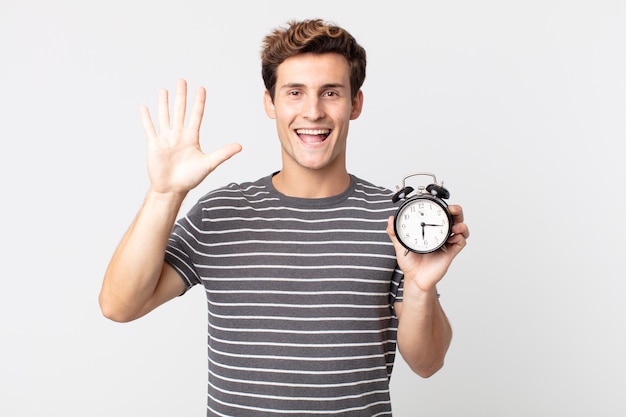 young handsome man smiling and looking friendly, showing number five and holding an alarm clock