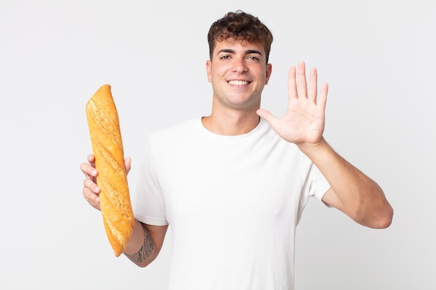 Young handsome man smiling and looking friendly, showing number five and holding a bread baguette