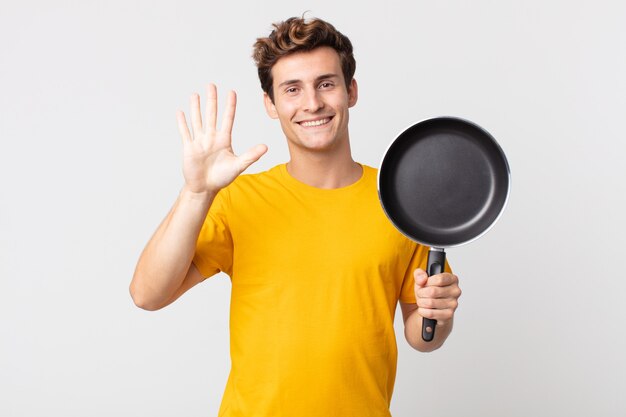 young handsome man smiling and looking friendly, showing number five and holding a cook pan