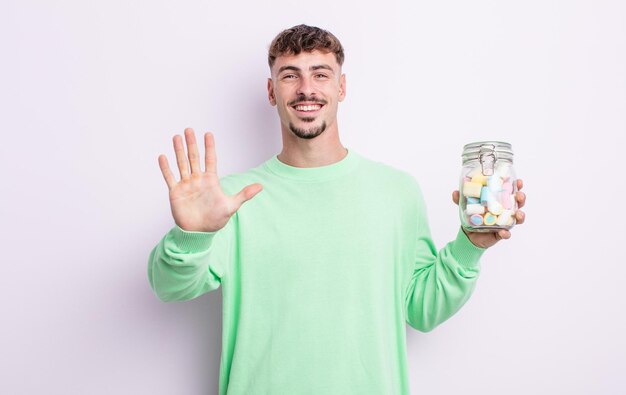 Young handsome man smiling and looking friendly, showing number five. jelly candies concept