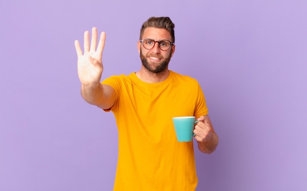 Giovane uomo bello che sorride e sembra amichevole, mostrando il numero quattro. e con in mano una tazza di caffè