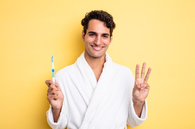 Young handsome man smiling and looking friendly, showing number three. bathrobe and toothbrush concept