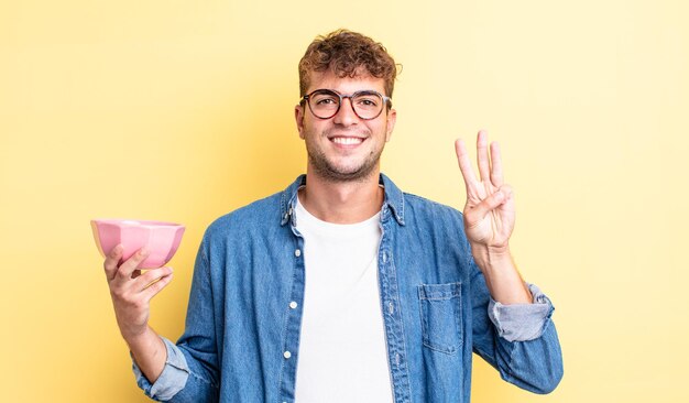 Giovane uomo bello che sorride e sembra amichevole, mostrando il numero tre. concetto di ciotola vuota