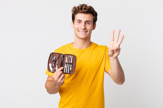 Young handsome man smiling and looking friendly, showing number three and holding a nails tools case