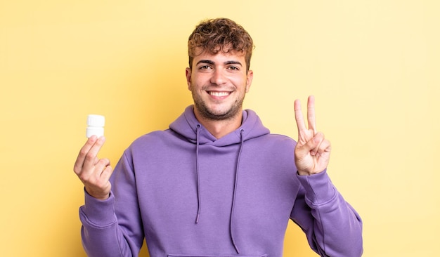 young handsome man smiling and looking friendly, showing number two. pills bottle concept