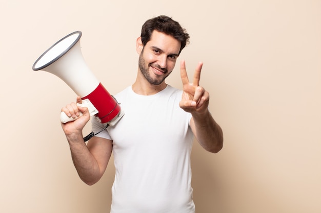 Young handsome man smiling and looking happy, carefree and positive, gesturing victory or peace with one hand