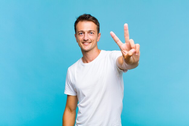 Young handsome man smiling and looking happy, carefree and positive, gesturing victory or peace with one hand