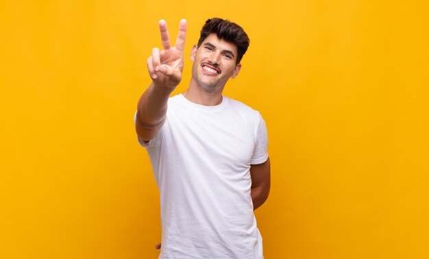 Young handsome man smiling and looking happy, carefree and positive, gesturing victory or peace with one hand