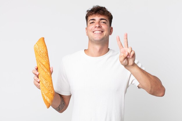 Young handsome man smiling and looking happy, gesturing victory or peace and holding a bread baguette