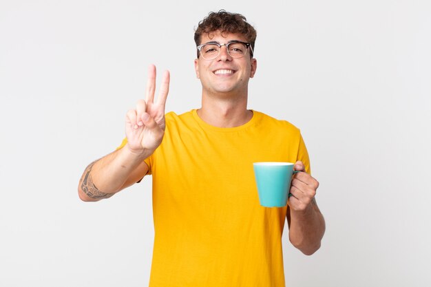 Young handsome man smiling and looking happy, gesturing victory or peace and holding a coffe cup