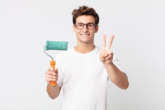 Young handsome man smiling and looking happy, gesturing victory or peace and holding a roller paint