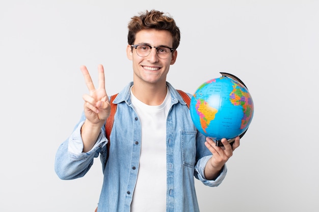 Giovane uomo bello che sorride e sembra felice, gesticolando vittoria o pace. studente con in mano una mappa del globo