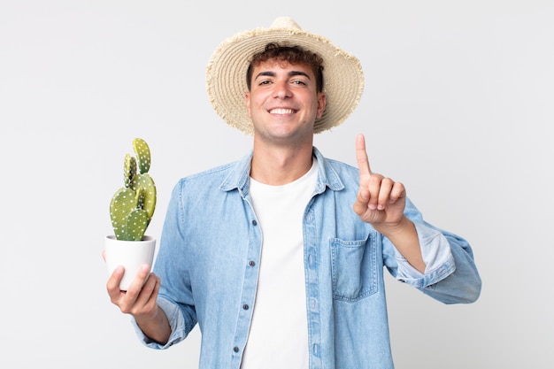 Giovane uomo bello che sorride con orgoglio e sicurezza facendo il numero uno. contadino con in mano un cactus decorativo